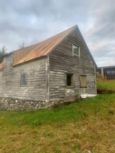 Fotografi av en gammel, gråtrekket bygning med steinmur og rustet tak sett fra siden.