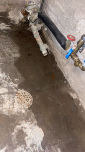 Fotografi av en teknisk installasjon i en kjele eller teknisk rom, som viser rørfittings, en svart isolert rørledning og et gulv med et avløpslokk.