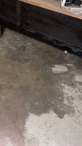 Fotografi av et gulv med synlige fukt- eller vannflekker, samt en betongvegg i bakgrunnen.