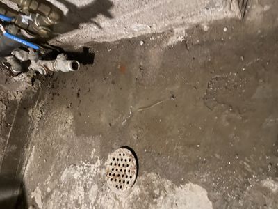 Fotografi av en våt gulvflate med en avløpsrør og rørledninger i hjørnet.