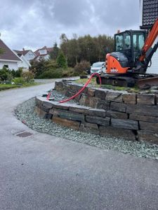 Fotografi av en byggeplass med en grøftemaskin, en steinmur og en vei.