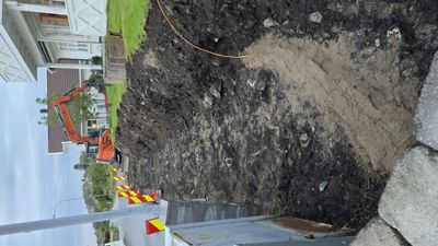 Fotografi av en byggeplass med en graverende maskin (graver) og utgravet jord. I bakgrunnen er det synlig en eksisterende bolig og en gate.