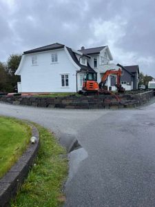 Fotografi av en hvit villa under bygging eller renovering, med en oransje graver i forgrunnen og en steinmur langs veien.