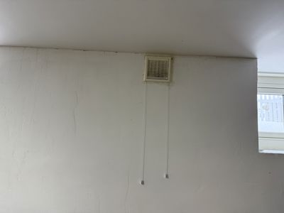 Fotografi av en hvit vegg med en ventilasjonsgitter og to kabler som henger ned.