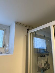 Fotografi av en innvendig vinkel i et bad, som viser en del av en dusjkabinett, en ventilasjonsskive i veggen og et vindu.