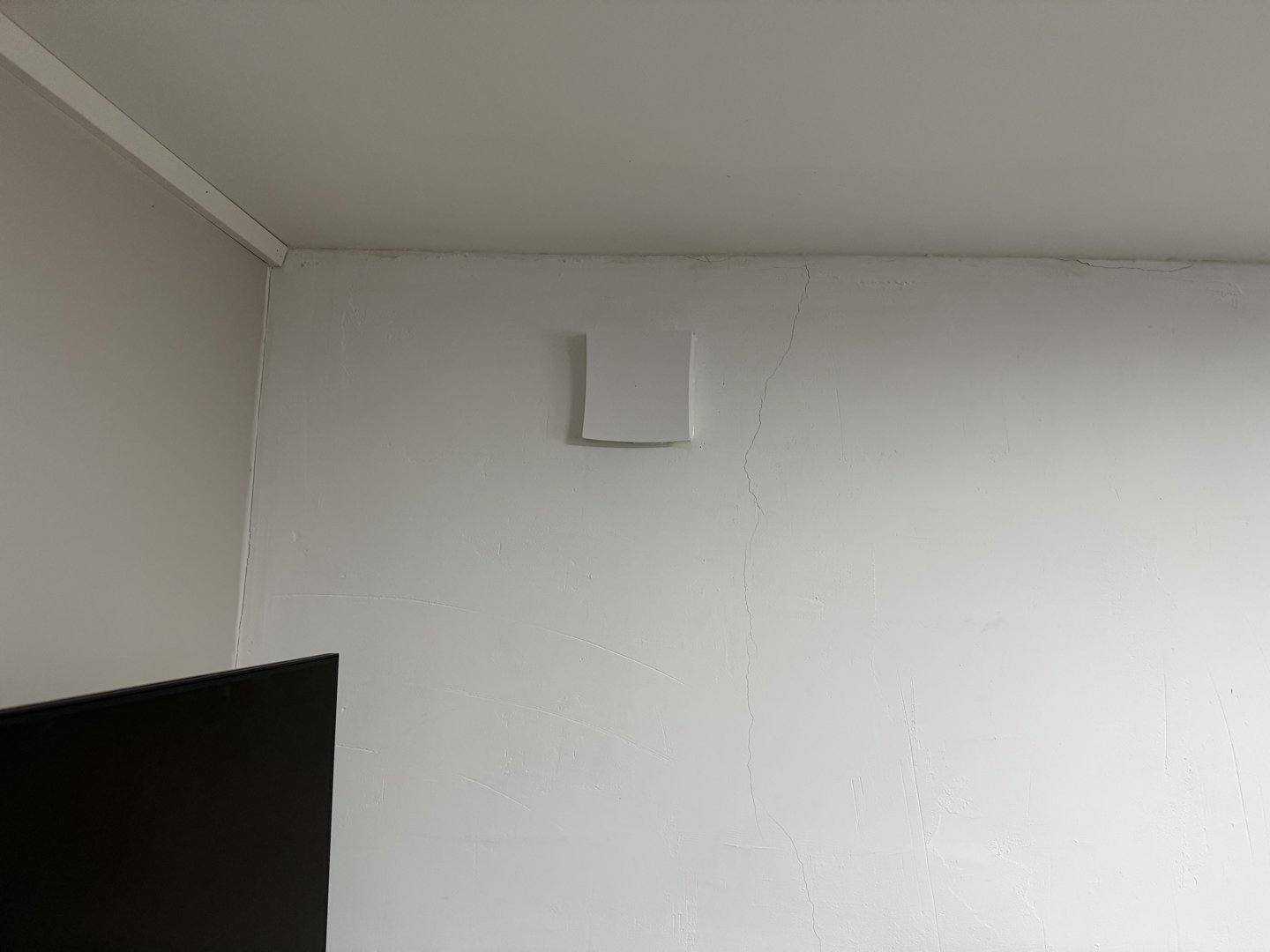 Fotografi av en hvit vegg i et rom, med en liten kvadratisk uttak i veggen og en del av en mørk TV-skjerm i forgrunnen.