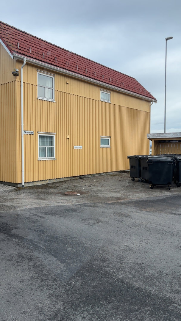 Fotografi av en gul bygning med rødt tak sett fra siden, med søppelcontainere i forgrunnen.