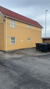 Fotografi av en gul bygning med rødt tak sett fra siden, med søppelcontainere i forgrunnen.