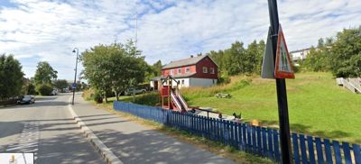 Fotografi av en vei, et barnehageanlegg med rødt hus og en blå hekk.