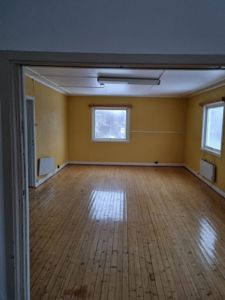 Fotografi av et tomt rom med gulmalt vegger, parkettgulv og vinduer sett gjennom en døråpning.