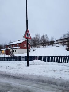 Fotografi av en snødekt gate med et hus, et barnehageanlegg og et trafikkskilt i forgrunnen.