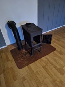 Fotografi av en stålkamin med røykledning i et rom med gulvlamell og panelvegger.