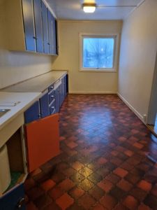 Fotografi av en tom kjøkkenrom med blå skap, hvite benker, rødt flisgulv og et vindu.