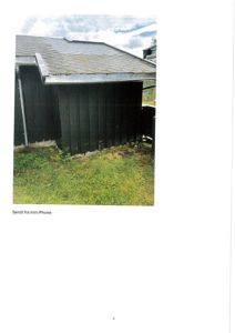 Fotografi av en mørk, trolig trepanelt bygning (kari eller uthus) sett fra siden. Bildet viser tak, vegger og grus/grass foran.