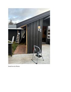 Fotografi av en mørk, trepanelt bygning (sannsynligvis en hytte eller uthus) sett fra siden. Bygningen har et skråtak, en åpen dør og en liten trappetrinn i forgrunnen. Det er også synlig en annen bygning i bakgrunnen.