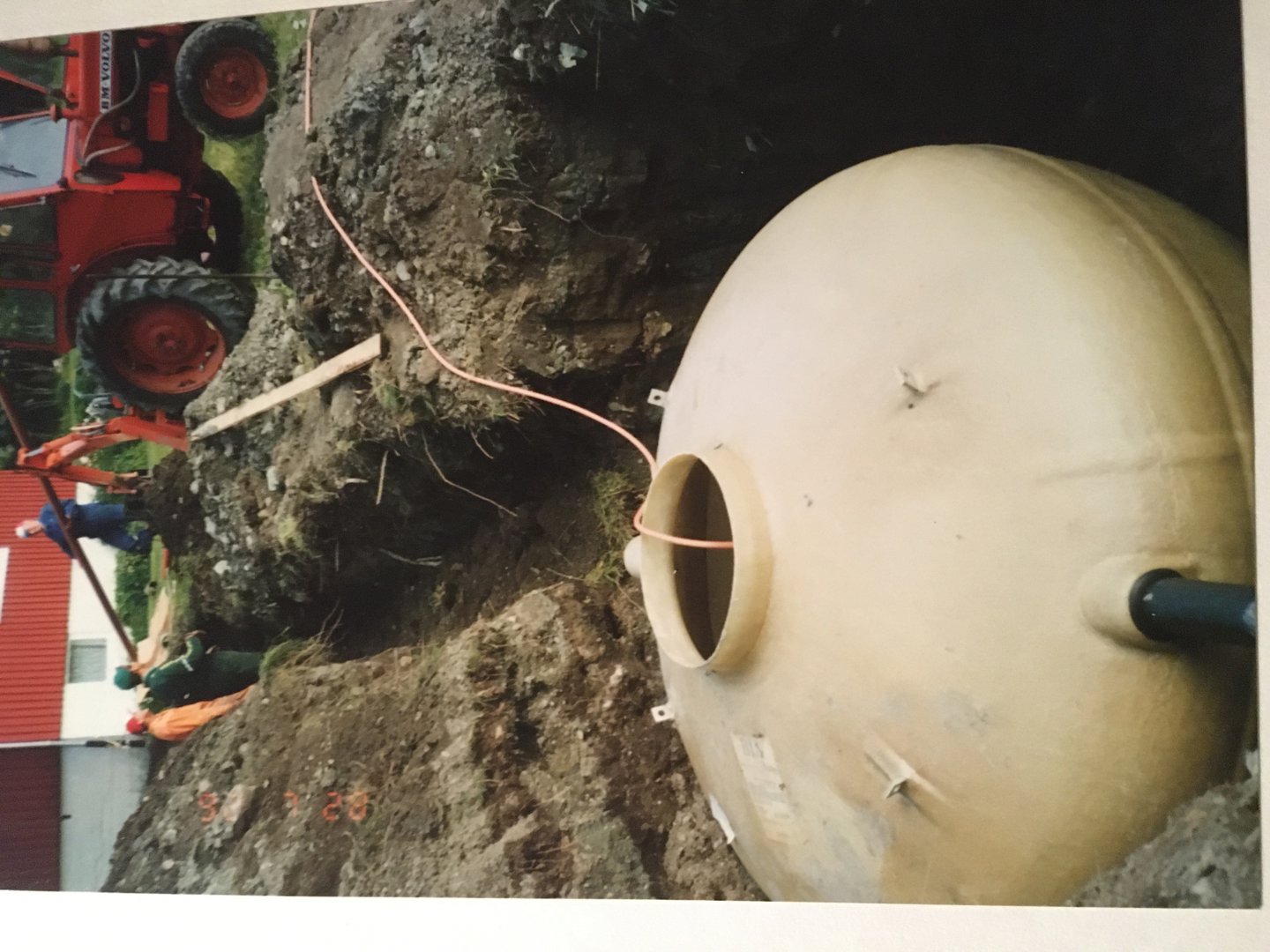 Fotografi av en stor, rund plasttank (sannsynligvis en septiktank eller lignende) som blir installert i en grøft ved hjelp av en rød lastebil. Bildet viser også arbeidere og en del av en bygning i bakgrunnen.