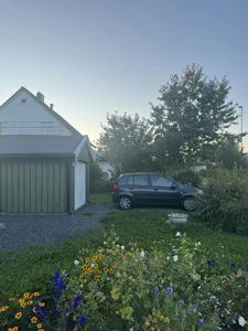 Fotografi av en grønn garasje og en hvit bygning sett fra hagen, med en bil parkert på grus.