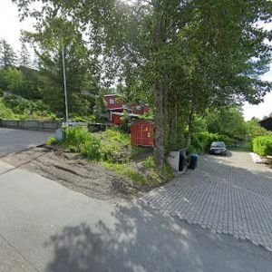 Fotografi av en tomt eller byggeplass med eksisterende beplantning, en rød container, og en bil parkert på en asfaltert overflate.