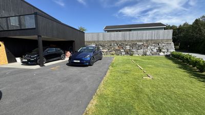 Fotografi av en moderne, mørk bygning med garasje, en blå bil og en grønn hage.