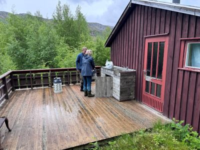 Fotografi av en rød trehytte med en uteplass, en vask og to personer.