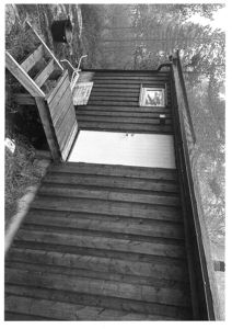 Fotografi av en liten, mørk trehytte sett fra en vinkel. Bildet viser fasaden med en hvit dør, et vindu, en takrenne og en trapp opp til en liten veranda.