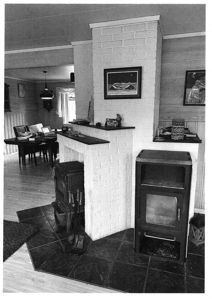 Sort-hvitt fotografi av et interiør med en steinovn, en peis og en spisebord i bakgrunnen.