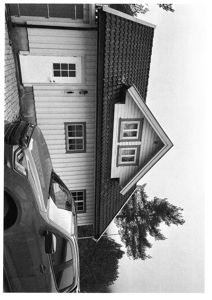 Sort-hvitt fotografi av en villa sett fra siden, med bil parkert foran og trær i bakgrunnen.