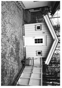 Sort-hvitt fotografi av en liten hytte eller uthus sett fra siden, med en hjulbar og en steinmur i forgrunnen.