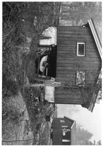 Sort-hvitt fotografi av en liten hytte eller uthus sett fra siden, med tilhørende terreng og vegetasjon.