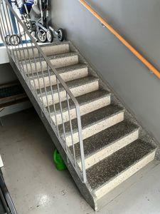 Fotografi av en trapp med terrazzo-trinn, metallhåndtak og en trelist langs veggen. I bakgrunnen er en barnevogn synlig.