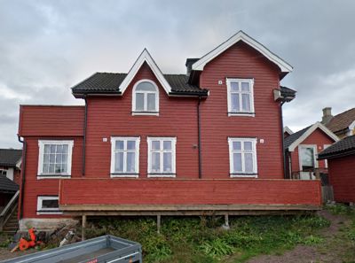Fotografi av en rød, to- eller treetasjers villa med hvite vinduer og et tak med dormer-vinduer. Bygningen står på en hevet grunnmur eller paller.