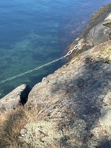 Fotografi av en kystlinje med klipper, hav og en ankerkjede som henger ned i vannet.