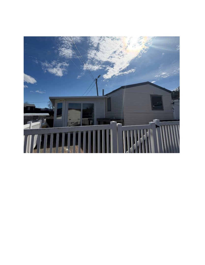 Fotografi av en hvit hytte eller hus sett fra bakgård eller side, med en hvit hekk i forgrunnen og sollyst i himmelen.