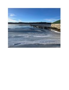 Fotografi av en islagt vannflate med en kai og en steinmur i forgrunnen, samt en bakke i bakgrunnen.