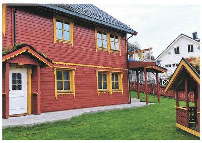 Fotografi av en rød, toetasers bygning med gule vindusrammer og et grønt tak. Bildet viser fasaden, en del av hagen og en tilstøtende hvit bygning.