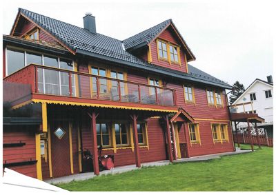 Fotografi av en rød, toetasjes trehytte med gul innramming, balkong og veranda.