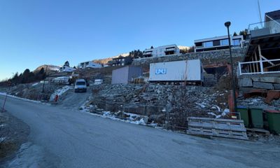 Fotografi av et byggefelt med moderne boliger i bakgrunnen, en vei i forgrunnen og byggemateriale (container, paller) på en steinete skråning.