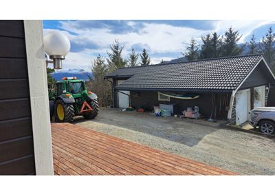 Fotografi av en eksisterende bygning (garasje/utleiebolig) sett fra en terrasse, med traktor og bil i forgrunnen.