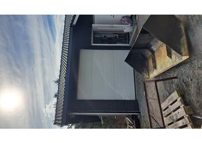 Fotografi av en bygning sett fra utsiden, med en stor hvit garasjeport og mørke fasadevegger. Bildet viser også solen, himmelen og gjenstander på bakken.