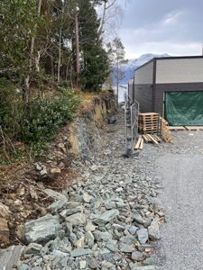 Fotografi av en byggeplass eller tomt ved vannet, med en nybygd eller renovert bygning i bakgrunnen. Bildet viser terrenget, steiner, palletter og en del av fasaden.