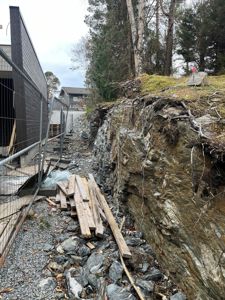Fotografi av en byggeplass som viser en steil, utgravet jordvegg (kanskje en grøft eller utgravning for fundament) ved siden av en nybygd eller renovert bygning med mørk fasade. Det er også synlig med materialer som tømmer og gjerde.