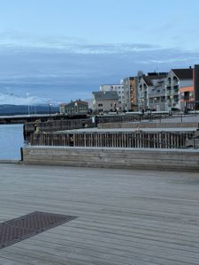 Fotografi av en havneområde med kai, båtplasser og flere bygninger i bakgrunnen mot en fjellkjede.