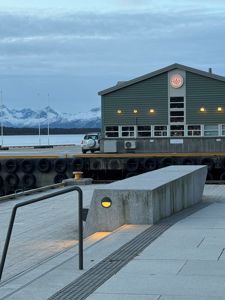 Fotografi av en grå bygning ved et kaiområde med snødekte fjell i bakgrunnen.
