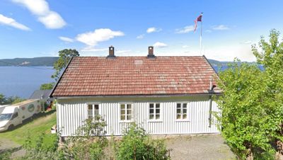 Fotografi av en hvit trehus med rødt tak, sett fra en viss avstand med vann og fjell i bakgrunnen.