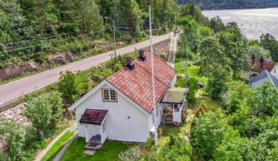 Et luftfoto av en hvit hytte med rødt tak, plassert ved en vei med skog og vann i bakgrunnen.