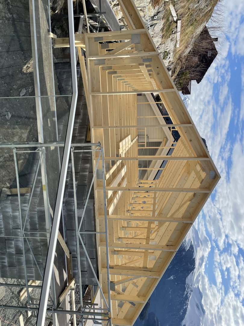 Fotografi av en byggeplass som viser en ferdigstilt takkonstruksjon i tre med stålskelett, sett fra en vinkel mot en snødekt fjellbakke.
