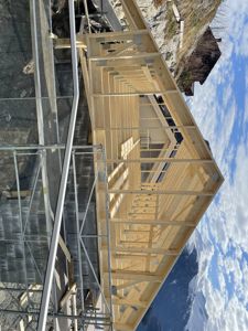 Fotografi av en byggeplass som viser en ferdigstilt takkonstruksjon i tre med stålskelett, sett fra en vinkel mot en snødekt fjellbakke.