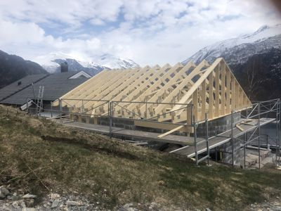 Fotografi av en bygning under oppføring med oppstilt takramme (trebjelker) og stålkonstruksjon, med fjell i bakgrunnen.