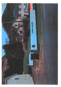 Fotografi av en eksisterende bygning (hvit, lang bygning med tekst på veggen) sett fra siden, med en vei og bakke i bakgrunnen.