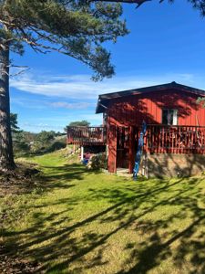 Fotografi av en rød hytte sett fra siden, med grønt gress og trær i forgrunnen.